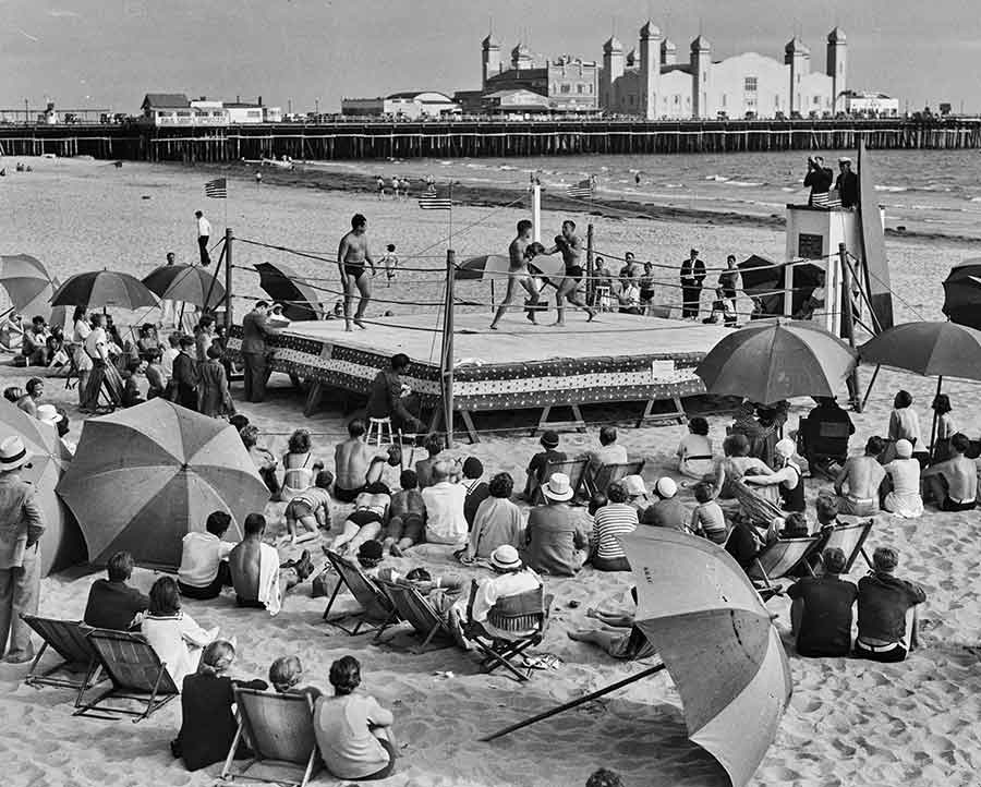 Boxers fighting in a ring in front of the Santa Monica Athletic Club, ca. 1934. The La Monica Ballroom is prominent on the Santa Monica Pleasure Pier in the distance. Photograph by Powell Press Service.