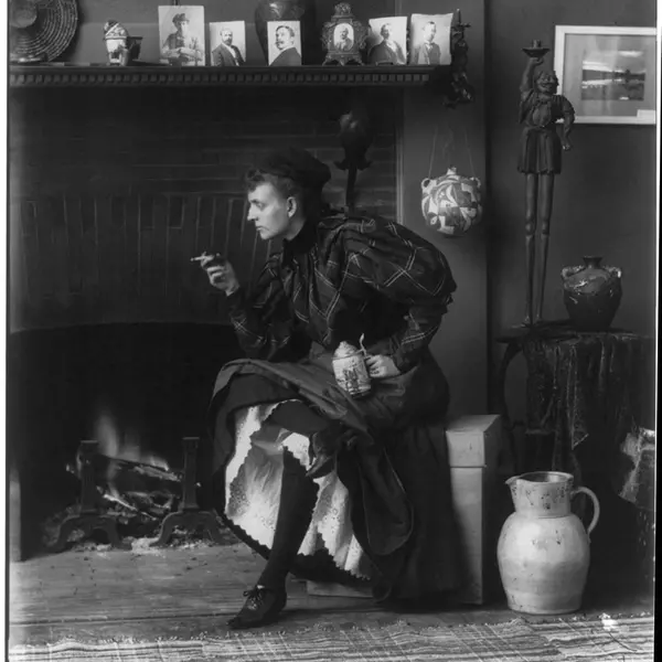 Black-and-white photo of Frances B. Johnston sitting in front of a fireplace, looking off-camera with a cigarette in her hand.