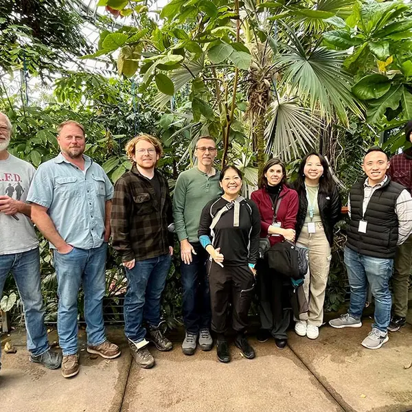 A group of people pose for a picture, standing in front of tropical trees.