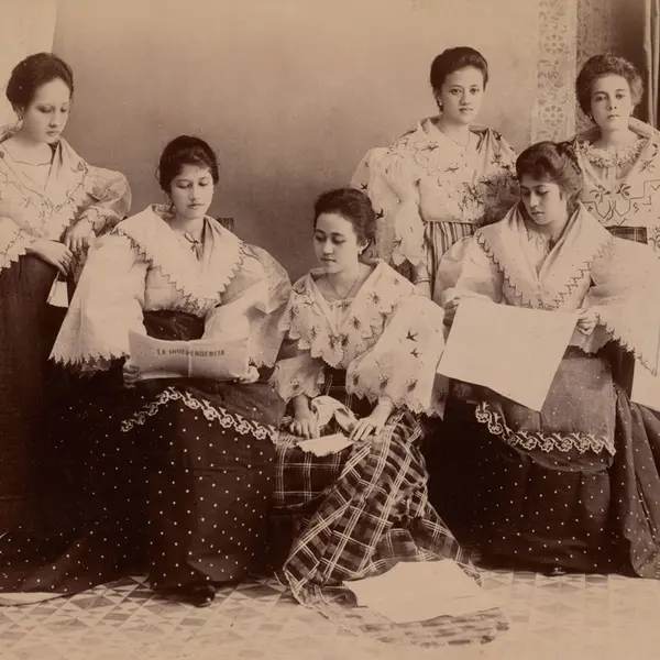 A group of six women in dresses pose while reading a newspaper.