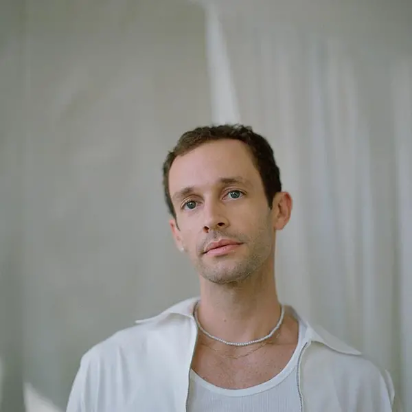 A person in a white undershirt and unzipped white jacket stares just off camera.