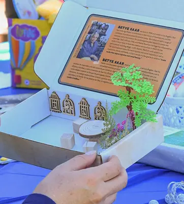 A person holds a small box with a lid, inside there is text on an orange background and objects glued in a diorama.
