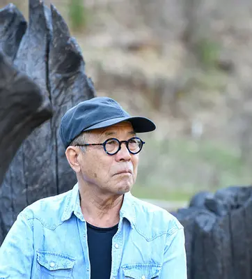 A man in a blue shirt and hat looks off camera, in front of a rock or tree formation.