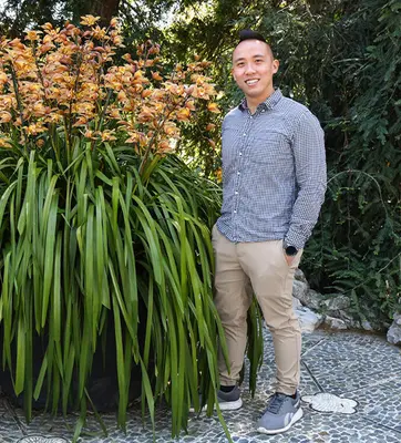 A smiling person stands near a large potted orchid with orange flowers.