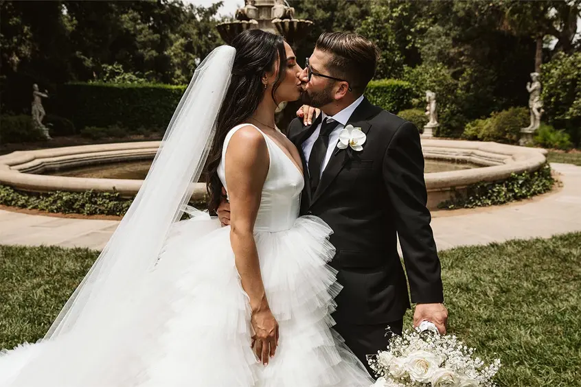 A wedding couple kiss in front of a large garden fountain.