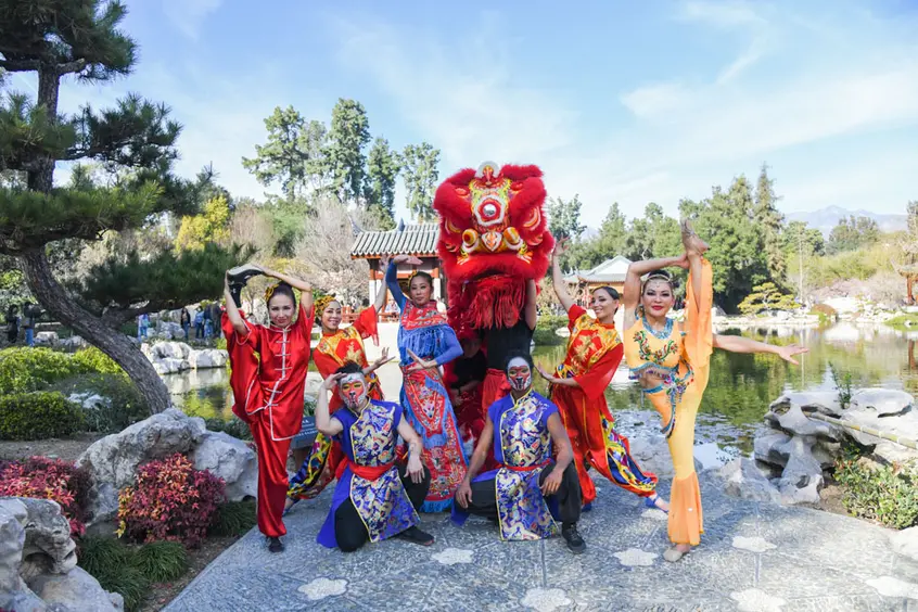 A group of performers in colorful outfits pose outdoors in a garden.