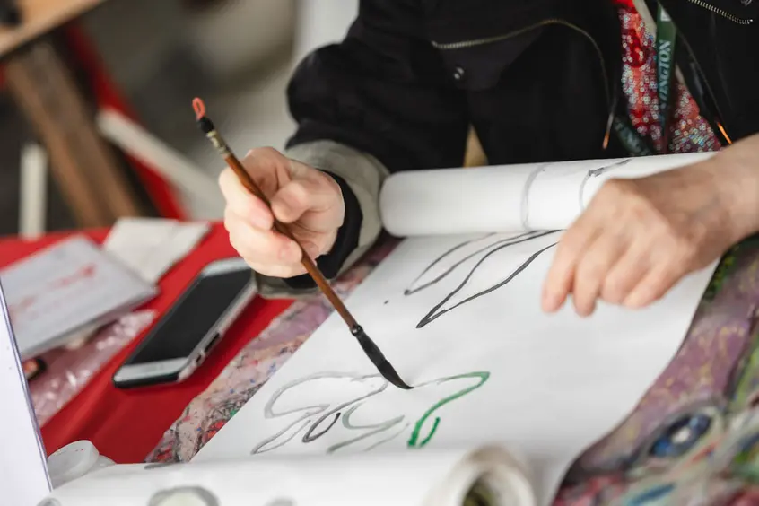 A person paints Chinese characters on white paper with a long wooden brush.