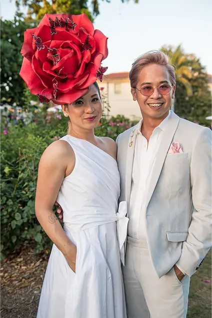 A couple in white, one wears a beautiful red rose hat with dancing butterflies.