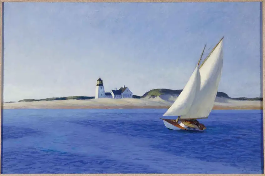 A leaning sailboat sits in calm deep-blue waters not far from a sandy beach and lighthouse.