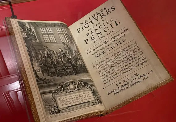 An open book in a glass case. On the left, page an illusration of a room of people. On the right, text reads "Natures Pictures Drawn by Fancies Pencil to the Life."
