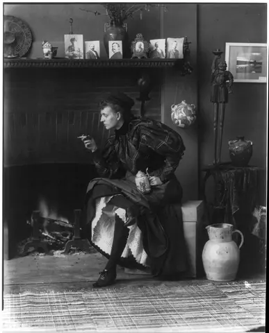 Black and white photo of a woman sitting on box in front of fireplace looking off-camera with cigarette in hand.