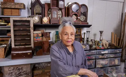 Color photograph of an older Black woman with white and grey hair pulled up to the top of her head in a short, curly ponytail. She is wearing a loose grey shirt and is standing in her artist studio, surrounded by wooden clocks, shelves, candlestick holders, and other miscellaneous items.