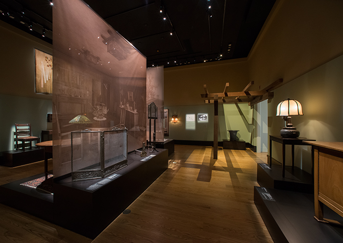 An image of the dining room of the Thorsen house appears on the left with examples of the Greene’s metalwork in front of it. In the background is the recreated fragment of a pergola from the 1903 Arturo and Helen Bandini house.