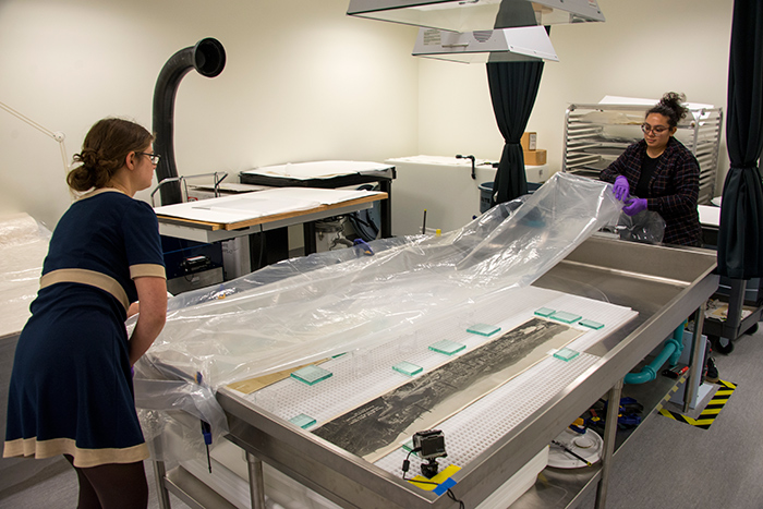 Intern Nicole Alvarado (right) and paper conservator Jessamy Gloor get ready to transfer a flattened photo into a drying stack. The time-lapse camera can be seen in the lower right corner of the humidification sink. Photo by Kate Lain.
